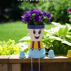 May include: A cheerful, cartoon-style planter with a smiling face, blue overalls, and yellow braces. The planter is decorated with white polka dots and has a purple flower pot with purple flowers on top. The planter has blue legs and is sitting on a wooden surface.