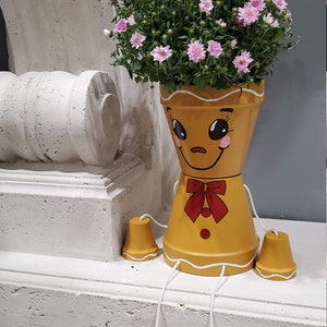 May include: A yellow gingerbread man planter with a white icing trim and a red bow. The planter has a happy face and is holding pink flowers. The planter is sitting on a white surface with four yellow legs.