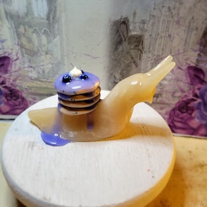 May include: A small, beige ceramic snail figurine carrying a stack of purple and white mini pancakes with blueberries on top.