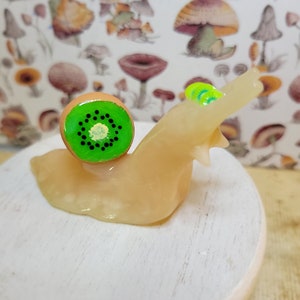 May include: A small, hand-painted ceramic snail sculpture with a green kiwi fruit shell. The snail is sitting on a white wooden base.