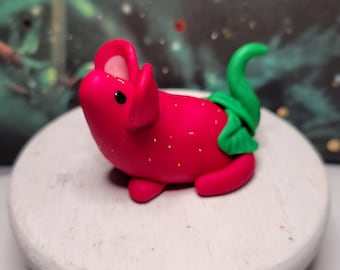 May include: A small, red, strawberry-shaped figurine with a green leaf tail and a small, white face. The figurine is made of polymer clay and has a glossy finish.