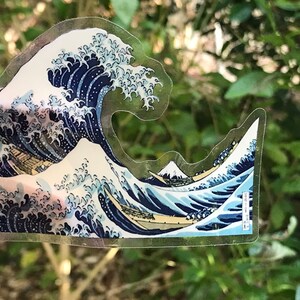 Pu&ograve; includere: Un adesivo trasparente con un'immagine blu e bianca della Grande Onda di Kanagawa di Hokusai. L'adesivo &egrave; su uno sfondo verde.