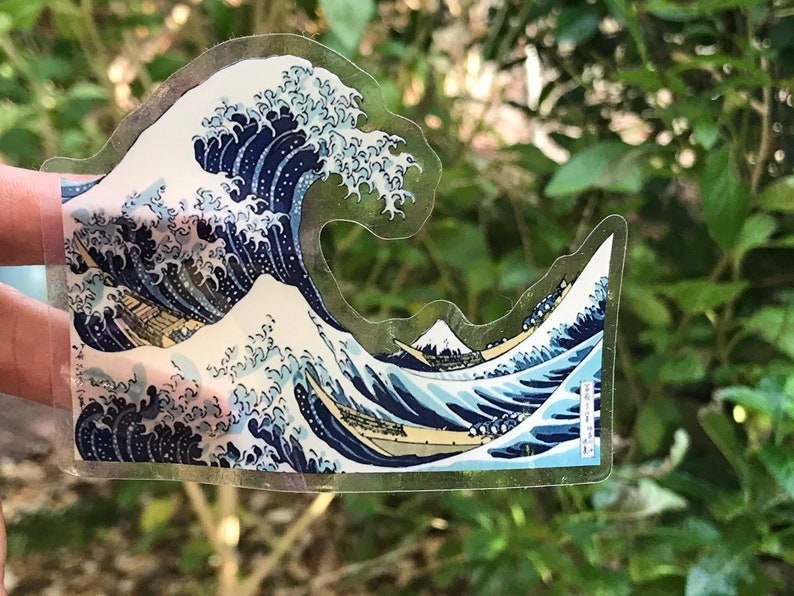 Pu&ograve; includere: Un adesivo trasparente con un'immagine blu e bianca della Grande Onda di Kanagawa di Hokusai. L'adesivo &egrave; su uno sfondo verde.