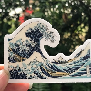 Pu&ograve; includere: Un adesivo con un'illustrazione dettagliata di un'onda gigante, ispirata alla famosa stampa giapponese su legno "La Grande Onda di Kanagawa" di Katsushika Hokusai. L'onda &egrave; rappresentata in tonalit&agrave; di blu e bianco, con una barca tradizionale giapponese e il monte Fuji sullo sfondo.