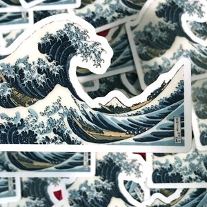 Pu&ograve; includere: Un adesivo con un'illustrazione blu e bianca di un'onda gigante che si infrange contro una montagna. L'onda &egrave; raffigurata in stile giapponese tradizionale, con una barca e persone che la cavalcano. L'adesivo &egrave; stampato su carta bianca e ha un bordo bianco.