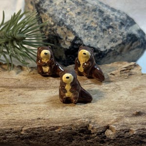 May include: Three small ceramic beaver figurines. Each beaver is brown with a cream-colored chest and face. The beavers are posed on a piece of driftwood. The background includes a large rock and a sprig of greenery.