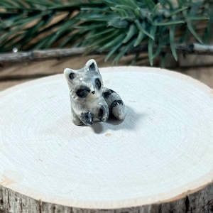 May include: A miniature ceramic raccoon figurine featuring a glossy white and grey finish with black markings around the eyes, ears, and on its striped tail. The small animal is depicted in a seated position with its front paws raised forward.