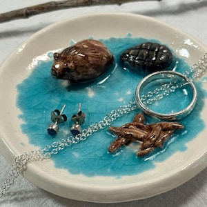 May include: A round white ceramic trinket dish featuring a central pool of crackled turquoise glaze. The dish contains a small brown sculpted beaver, a dark textured stone, a silver chain necklace with a ring pendant, and two blue stud earrings.