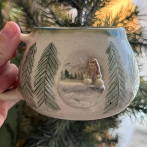 May include: Cream-colored ceramic mug with a forest scene featuring a Bigfoot figure. The mug has a handle and is decorated with hand-painted pine trees and a mountain landscape. The rim is a darker green color.