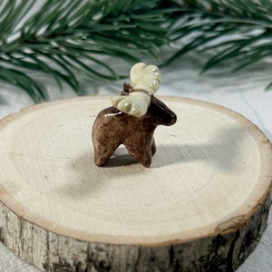 May include: A small, hand-sculpted ceramic figurine of a moose with a glossy, mottled brown body and smooth, cream-colored antlers. The moose stands in profile, showcasing a rounded form and distinct, curved antlers positioned atop its head.
