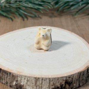 May include: A small, cream-colored ceramic figurine of a bear with a smooth, glossy finish. The bear has simple, dark-painted eyes and a rounded snout, depicted in a seated position with its front paws resting against its chest.