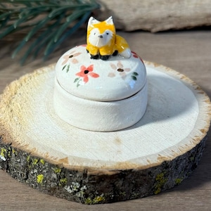 Peut inclure: Petite boîte à bijoux ronde en céramique blanc cassé, avec un couvercle orné de fleurs roses et pêche. Une figurine de renard jaune et orange est posée sur le couvercle. La boîte est sur une tranche de bois.