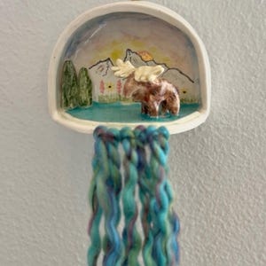 May include: A white ceramic wall hanging featuring a semi-circular bowl containing a hand-painted scene of a brown moose standing in blue water before mountains, trees, and flowers, with long, wavy teal and purple yarn tassels hanging below.