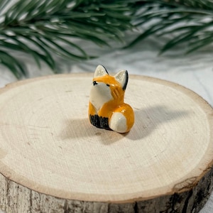 Pode incluir: Uma pequena estatueta de raposa em cerâmica com uma pelagem laranja brilhante, peito e rosto brancos, e detalhes pretos nas orelhas e patas. A raposa está representada numa posição sentada com a cabeça inclinada para cima e acabamento liso.