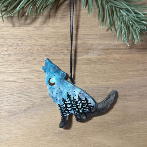 May include: A ceramic wolf-shaped ornament, painted with a night sky design. It features a crescent moon, stars, and a silhouette of trees. The ornament hangs from a black string. The colour palette includes blues, blacks, and browns.