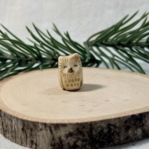 May include: A small, cream-colored ceramic owl figurine featuring etched feather details, closed eyes, and a metallic gold-painted beak. The owl has a rounded body shape with a textured surface and a subtle, neutral color palette throughout.