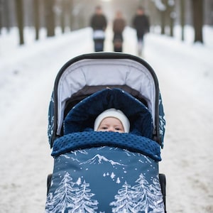 May include: A baby in a stroller on a snowy path. The stroller has a navy blue cover with a white mountain and tree design. The baby is wearing a white hat and is bundled in a navy blue blanket.