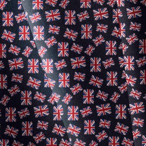 May include: A dark navy blue fabric featuring a repeating pattern of small Union Jack flags in red, white, and blue. The flags are oriented in various directions across the surface, creating a dense, uniform graphic print on the textile material.