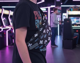 Puede incluir: Una riñonera negra con un patrón repetitivo de mandos de videojuegos y botones direccionales en blanco y azul claro. El bolso cuenta con una cremallera negra y una etiqueta rectangular de tela gris en la esquina frontal inferior.
