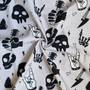 May include: A light gray fabric featuring a repeating black graphic print of skulls, electric guitars, lightning bolts, small bones, and skeletal hands making rock and roll gestures. The pattern is distributed across the surface of the textile.