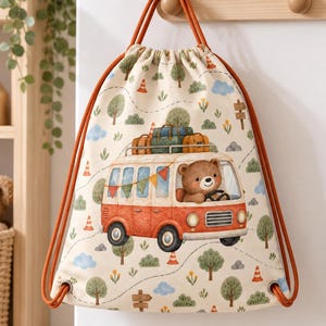 May include: A cream-colored drawstring backpack featuring a repeating pattern of trees, clouds, and orange traffic cones. The central graphic depicts a brown bear driving an orange camper van with luggage on the roof and colorful bunting.