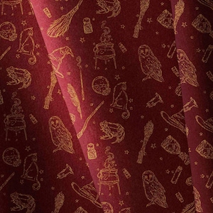 Può includere: Un tessuto bordeaux scuro con un motivo ripetuto a linee dorate raffigurante elementi magici tra cui gufi, gatti, rane, calderoni fumanti, bacchette, scope, boccette di pozioni e piccole stelle sparse sulla superficie rosso scuro.