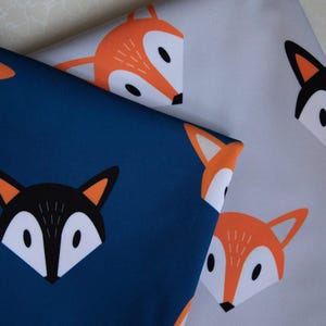 May include: Two pieces of fabric featuring a repeating fox face pattern. One piece is dark blue with black, white, and orange fox faces. The other piece is light gray with orange, white, and black fox faces. The fox faces have pointed ears.