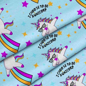 May include: Light blue fabric featuring a repeating pattern of white unicorns with multicolored manes and tails, wearing black sunglasses. The unicorns are performing a dab pose, accompanied by yellow and purple stars and the text I CHOOSE TO BE AUNICORN.