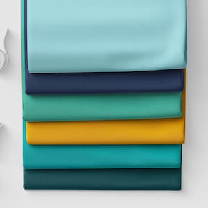 Op de afbeelding: Een stapel van zes gevouwen stoffen in verschillende effen kleuren, waaronder lichtblauw, marineblauw, taling, mosterdgeel, turkoois en donker bosgroen, gerangschikt in een verticale kolom met gladde, matte texturen en strakke randen.