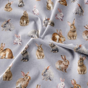 May include: A light grey fabric featuring a repeating pattern of watercolour-style rabbits in various shades of brown, tan, white, and grey. The bunnies are depicted in different poses, including sitting and resting, scattered across the textured surface.