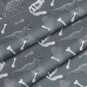 May include: Grey fabric featuring a repeating pattern of dinosaurs, dinosaur skulls, and bones. The dinosaurs are in shades of grey, with white bones and skulls. The fabric appears slightly folded.