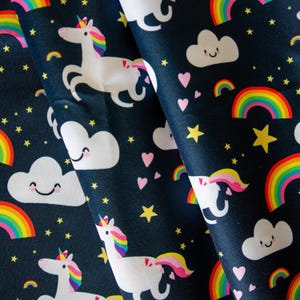 May include: A dark navy blue fabric featuring a repeating pattern of white unicorns with rainbow manes and tails, smiling white clouds, vibrant multi-colored rainbows, small yellow stars, and light pink hearts scattered across the surface.