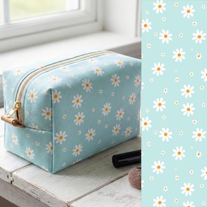May include: A light blue cosmetic bag with a daisy pattern. The bag has a gold zipper and a small leather pull tab. The daisy pattern is white with yellow centers. The bag is sitting on a white wooden surface.