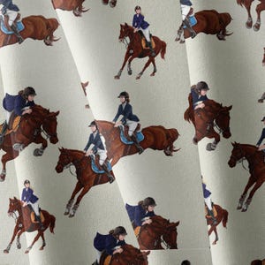 May include: A light-colored fabric featuring a repeating pattern of equestrian riders in navy blue jackets and helmets jumping over obstacles on brown horses. The riders are depicted in various positions, including mid-jump and galloping, with detailed saddles.