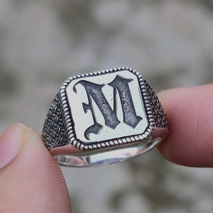 Puede incluir: Un anillo de sello plateado con la letra "M" en una fuente audaz y estilizada. El anillo tiene una cara rectangular con un borde texturizado y un acabado pulido.