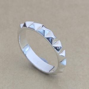 May include: A silver ring with a repeating geometric design of triangular shapes. The ring is made of a shiny, reflective metal and sits on a neutral-toned surface. The ring's design is modern and minimalist.