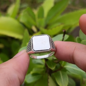 Può includere: Un anello d'argento con una faccia quadrata e vuota. L'anello ha una fascia dettagliata e strutturata e una superficie liscia e lucida. L'anello è tenuto su uno sfondo di foglie verdi.
