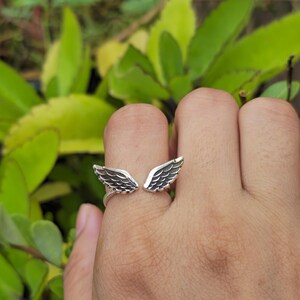 May include: A silver-colored ring featuring an open design with two detailed wing shapes. The wings are positioned to appear as if they are wrapping around the finger. The ring is shown on a hand against a backdrop of green foliage.