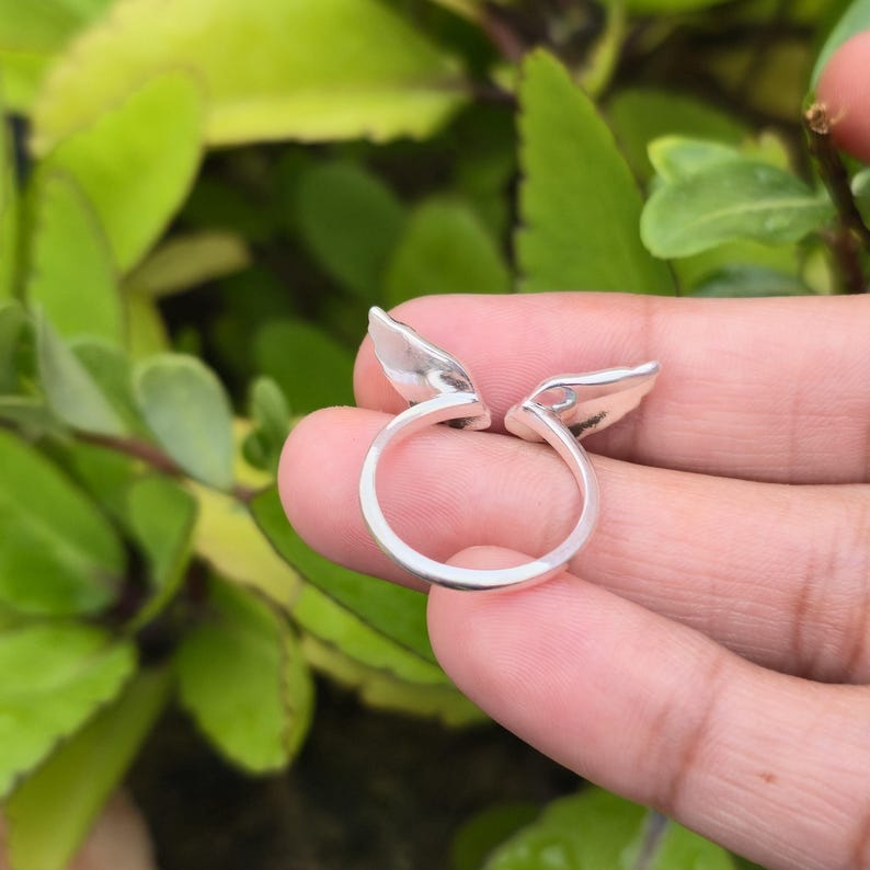 May include: A silver ring with a unique design featuring two stylized wing shapes. The ring is held in a hand against a backdrop of green foliage, highlighting the ring's polished surface and intricate details.