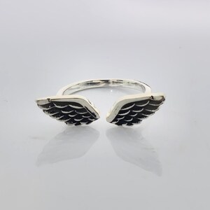 May include: A silver-colored ring featuring an open design with two detailed wing shapes. The wings are dark with intricate feather detailing, creating a contrast against the ring's shiny band. The ring is designed to be worn on a finger.