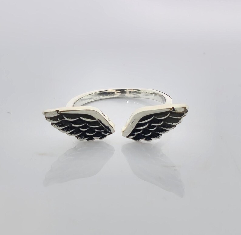 May include: A silver-colored ring featuring an open design with two detailed wing shapes. The wings are dark with intricate feather detailing, creating a contrast against the ring's shiny band. The ring is designed to be worn on a finger.