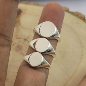 Puede incluir: Tres anillos de sello plateados con acabado pulido. Cada anillo presenta una cara ovalada lisa y una banda cónica. Los anillos se muestran en un dedo sobre un fondo de madera clara.