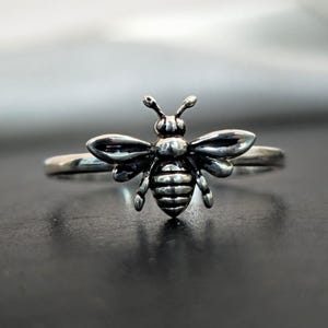 Può includere: Un anello argentato con un design dettagliato a forma di ape. L'ape ha ali e corpo strutturati, con antenne e una piccola testa. La fascia dell'anello è sottile e semplice, creando un delicato gioiello.