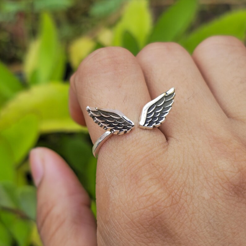 May include: A silver-colored ring featuring an open wing design. The wings are detailed with etched feather patterns. The ring is shown on a finger against a blurred green background, suggesting an outdoor setting. The ring is a piece of jewelry.