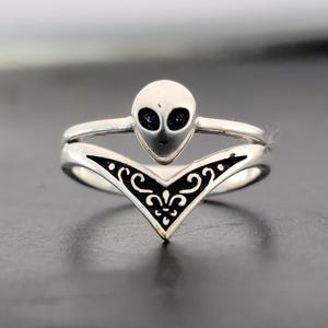 May include: A silver ring featuring an alien head design with black eyes. The ring has a V-shaped band with a black filigree pattern. The alien head is centered above the band.