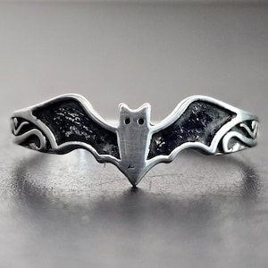 May include: A silver-toned ring featuring a bat design. The bat has outstretched wings with textured details and a central body with two small eye-like features. The ring's band has decorative scrollwork on each side of the bat.