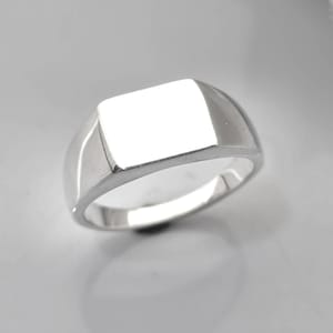 May include: A polished silver signet ring with a rectangular face. The ring has a smooth, rounded band and a bright, reflective surface. The design is simple and classic, suitable for everyday wear.