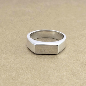 May include: A silver signet ring with a rectangular face and a geometric design. The ring is made of a shiny, metallic material and is set against a neutral background. The ring's design is simple and modern.