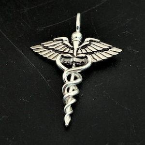 Può includere: Un ciondolo color argento raffigurante il simbolo del Caduceo, un bastone con due serpenti attorcigliati e ali nella parte superiore. Il ciondolo è probabilmente in metallo ed è progettato per essere indossato come gioiello.