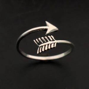 Può includere: Un anello color argento a forma di freccia. Il design della freccia avvolge il dito, con la punta della freccia rivolta verso l'esterno. L'anello è posto su uno sfondo scuro.
